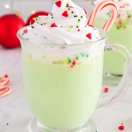 A green glass of peppermint hot chocolate topped with whipped cream, sprinkles, and a candy cane.
