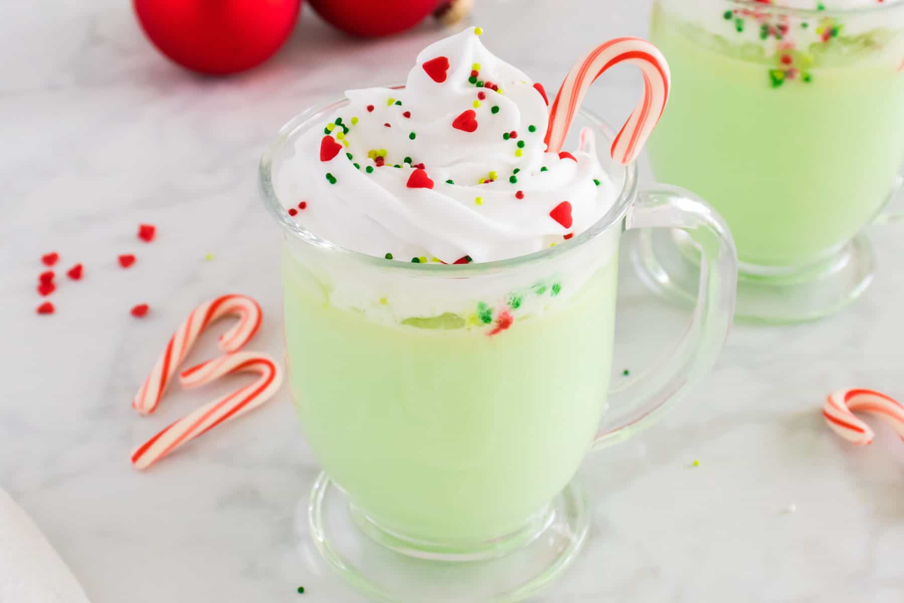 Green-colored peppermint white hot chocolate in a glass mug topped with whipped cream, sprinkles, and a candy cane.