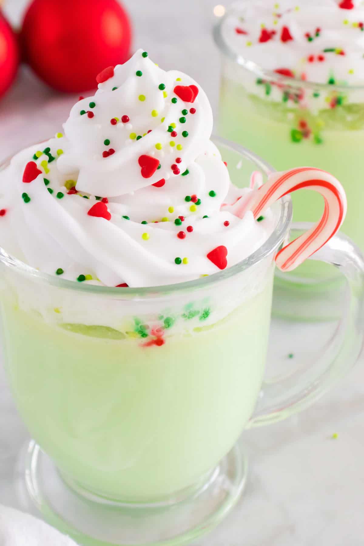 A closeup of a glass mug of green hot chocolate topped with whipped cream, festive sprinkles, and a candy cane.