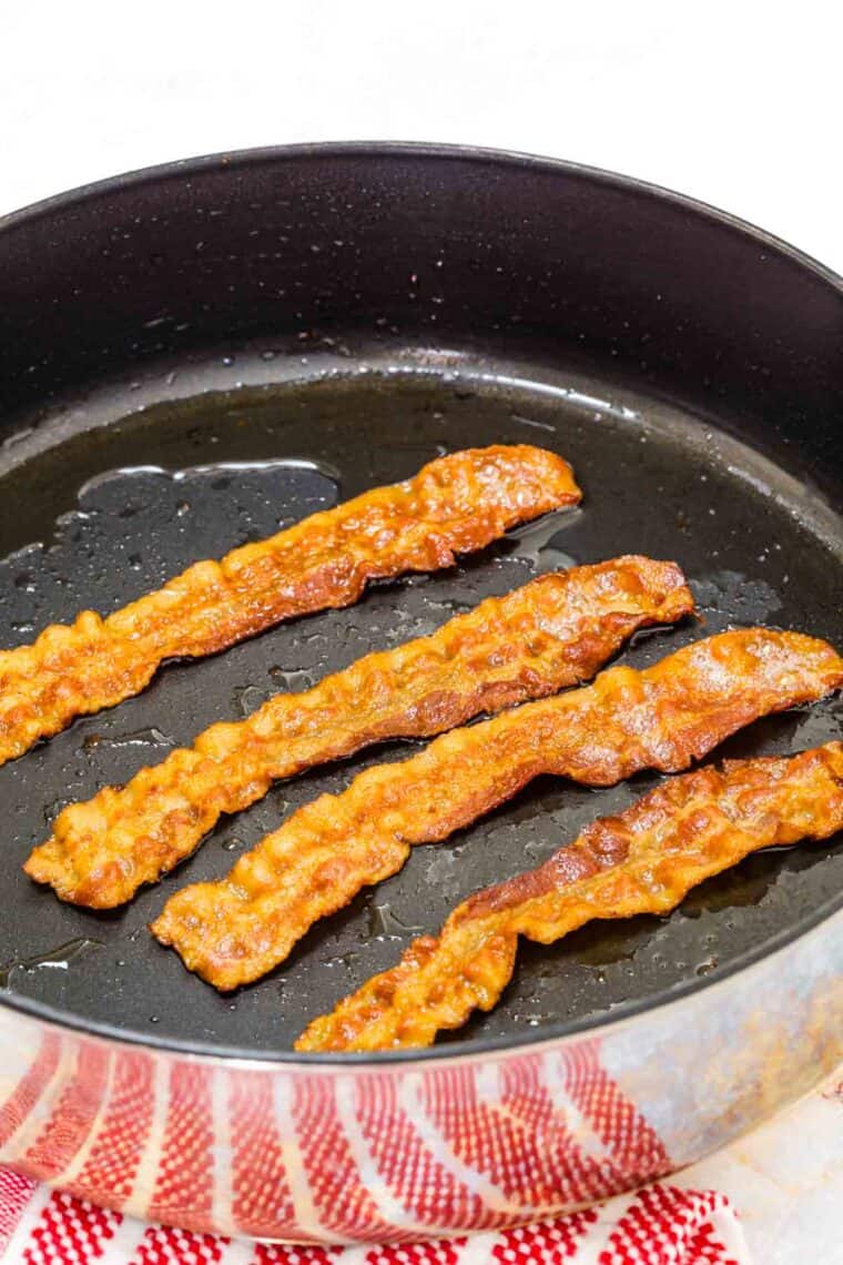 Four strips of bacon cook in a skillet.