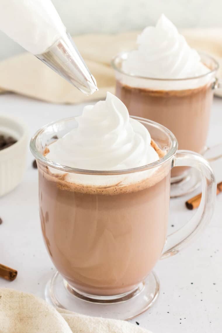 Whipped cream is piped onto glasses of hot chocolate.