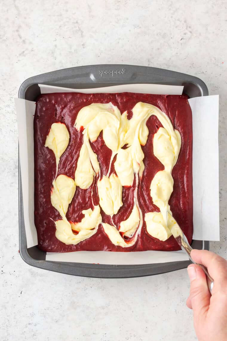 A knife swirls sweetened cream cheese into red velvet brownie batter.