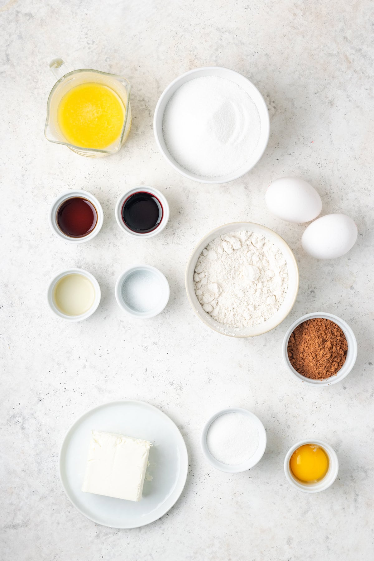Ingredients needed to make red velvet brownies are shown portioned out into bowls: vanilla, melted butter, sugar, eggs, egg yolk, gluten free flour, cocoa powder, red food coloring, sugar.