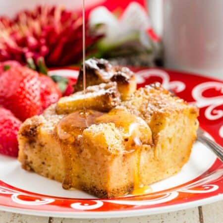 Syrup being poured on and dripping off of a piece of french toast casserole on a red and white plate.