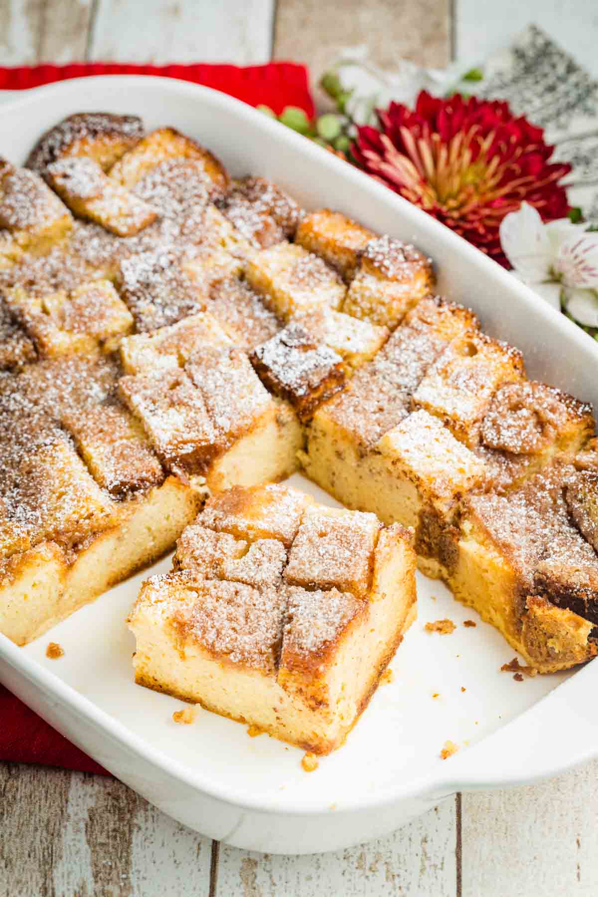 Pieces of French toast casserole are shown in a white baking dish surrounded by red and white flowers.
