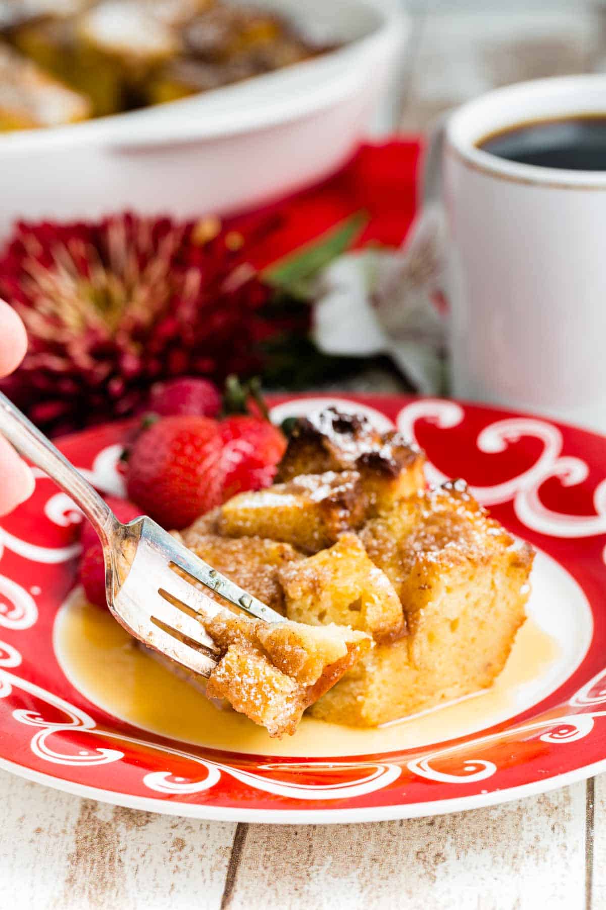 A fork digs into a piece of French toast casserole on a red and white plate. Strawberries and red flowers are seen in the background.