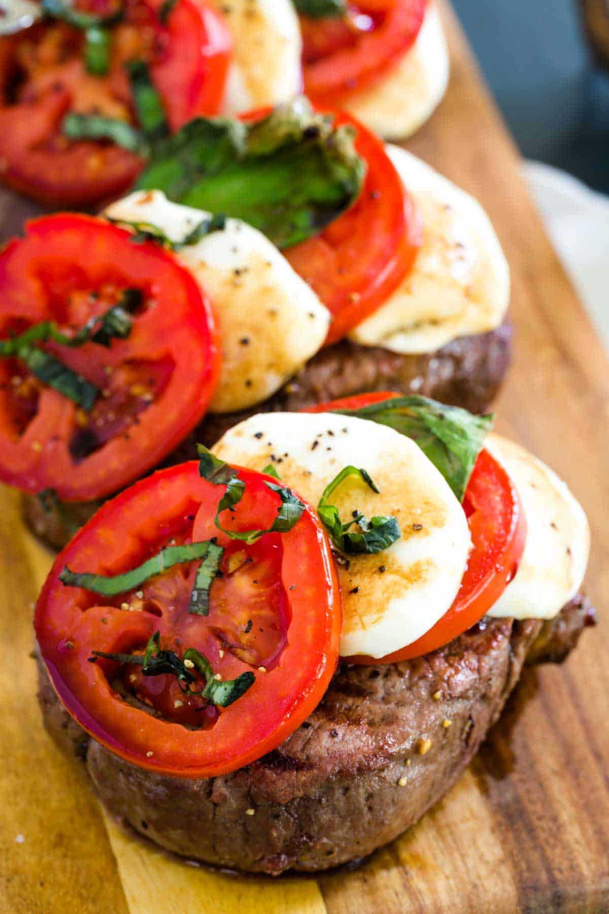 Grilled Caprese steaks are shown on a wooden board featuring slices of mozzarella and tomatoes.