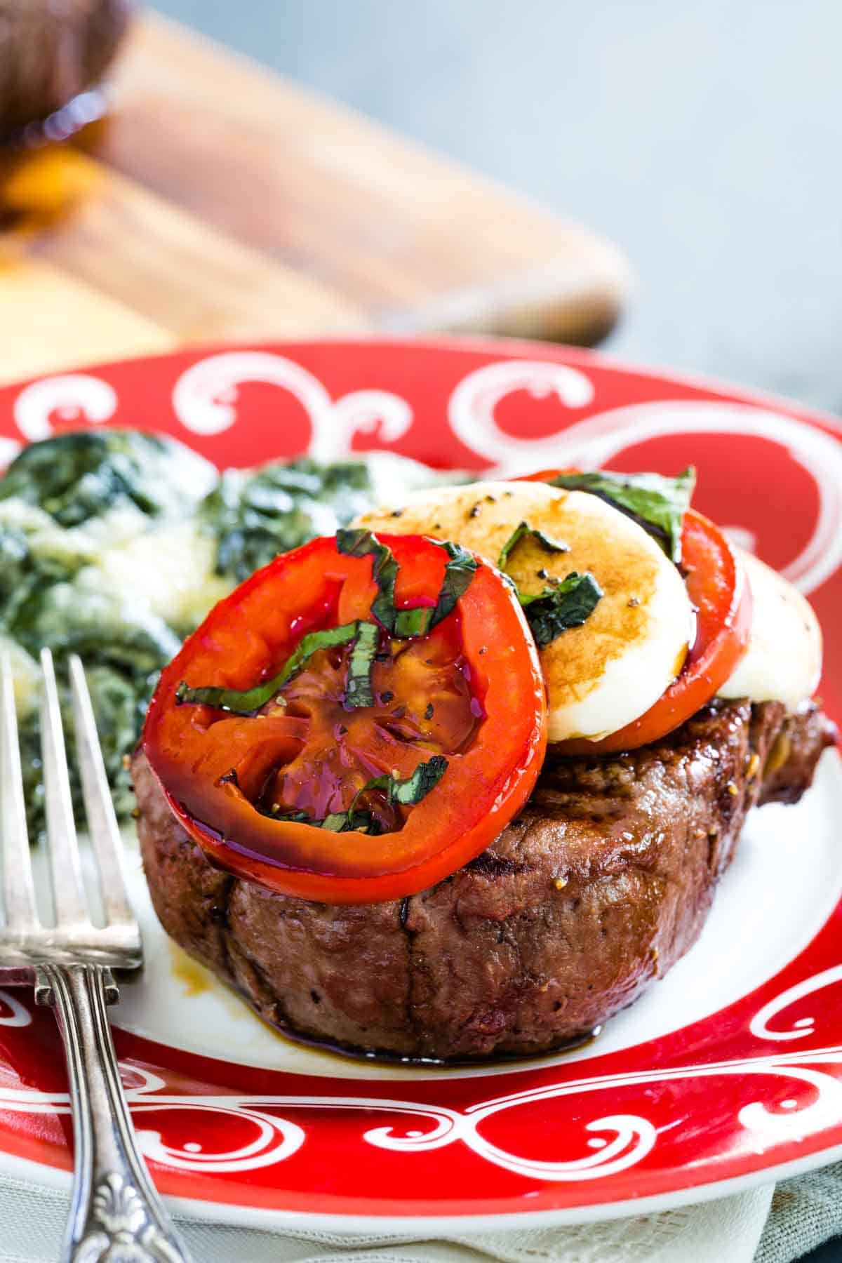 A Caprese steak, topped with basil, tomatoes, and mozzarella slices, is shown on a red and white plate with a fork to its left.