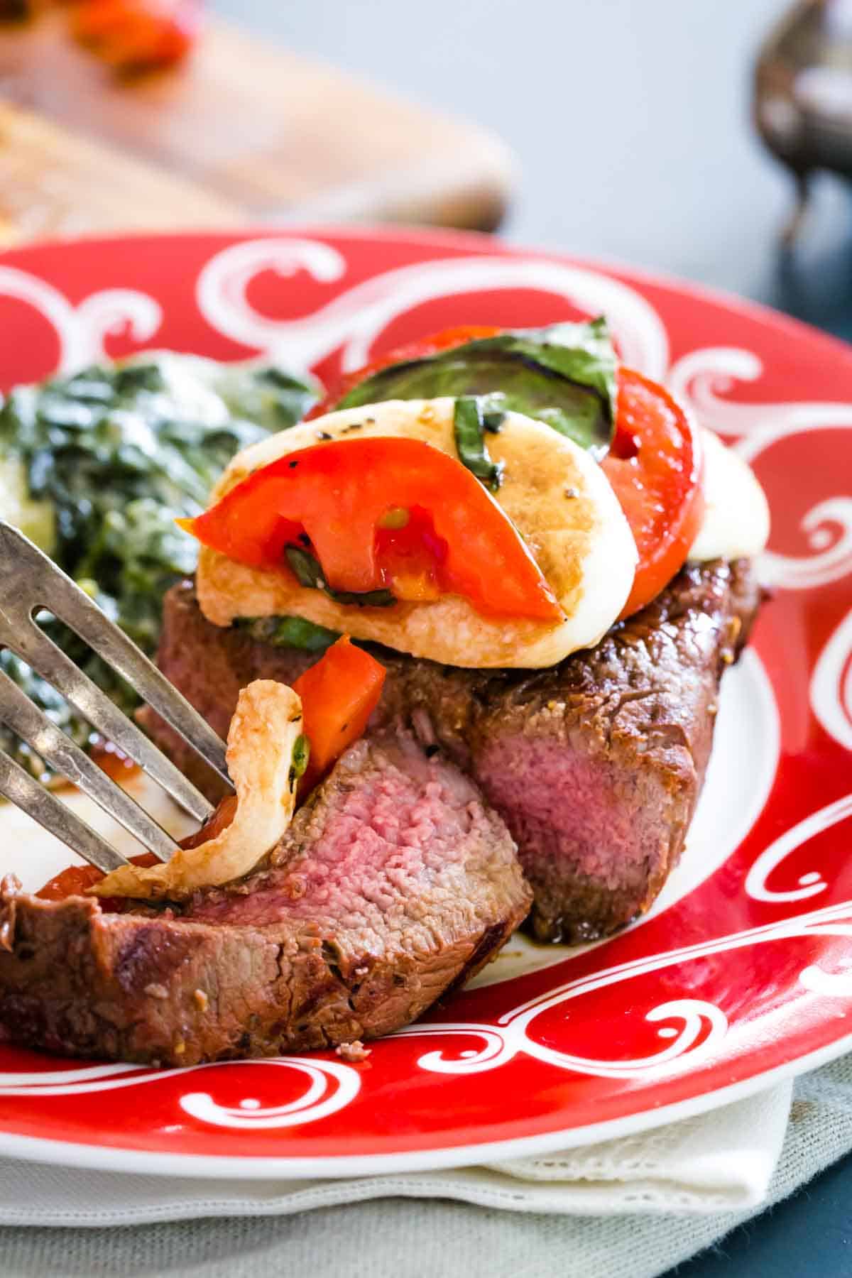 A fork picking up a slice of caprese filet mignon from a plate with the steak on it.