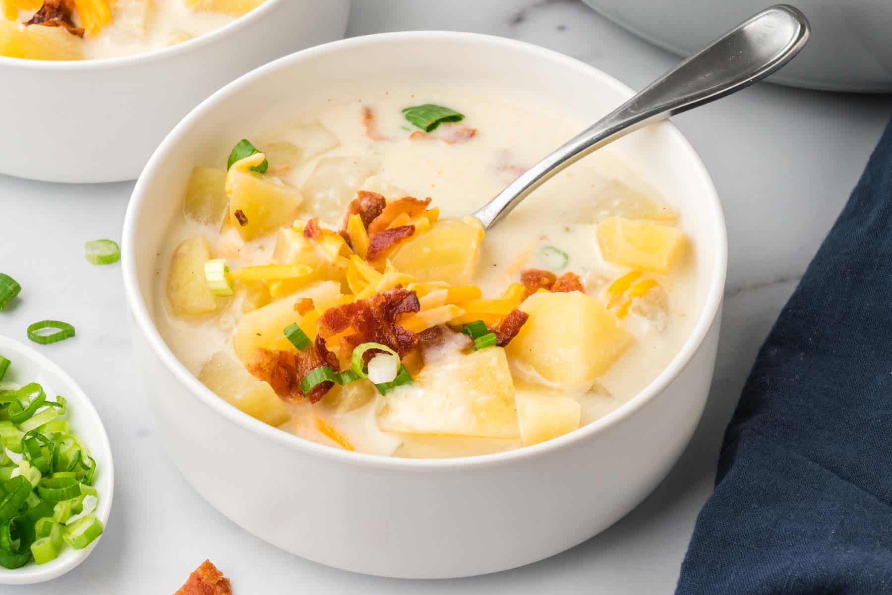 A white bowl holds cream of potato soup topped with colorful garnishes and a spoon is inserted into it.