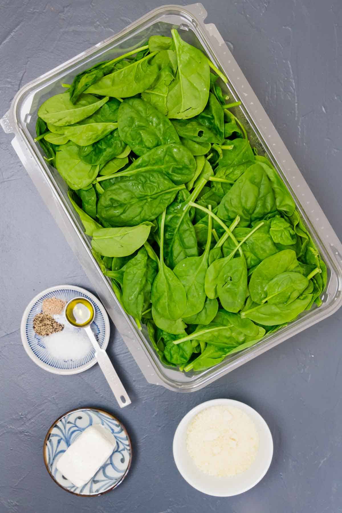 The ingredients needed to make creamed spinach are shown portioned out on a gray background: baby spinach leaves, cream cheese, parmesan cheese, salt and pepper, olive oil, and nutmeg.