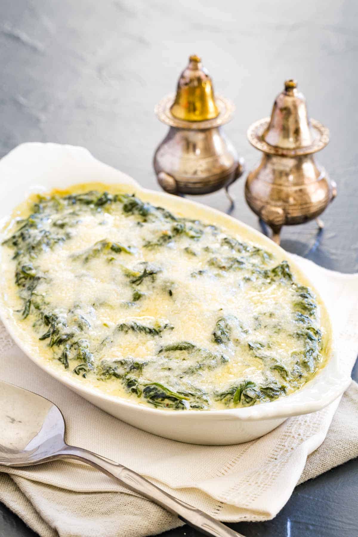 A white oval baking dish is shown full of Parmesan-topped creamed spinach, on a white napkin with serving utensils and metal salt and pepper shakers next to it.