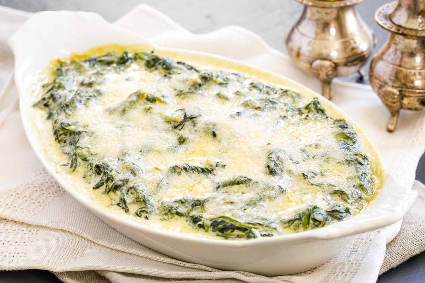 An oval white baking dish holds melted cheese-topped creamed spinach with metal salt and pepper shakers to its right.