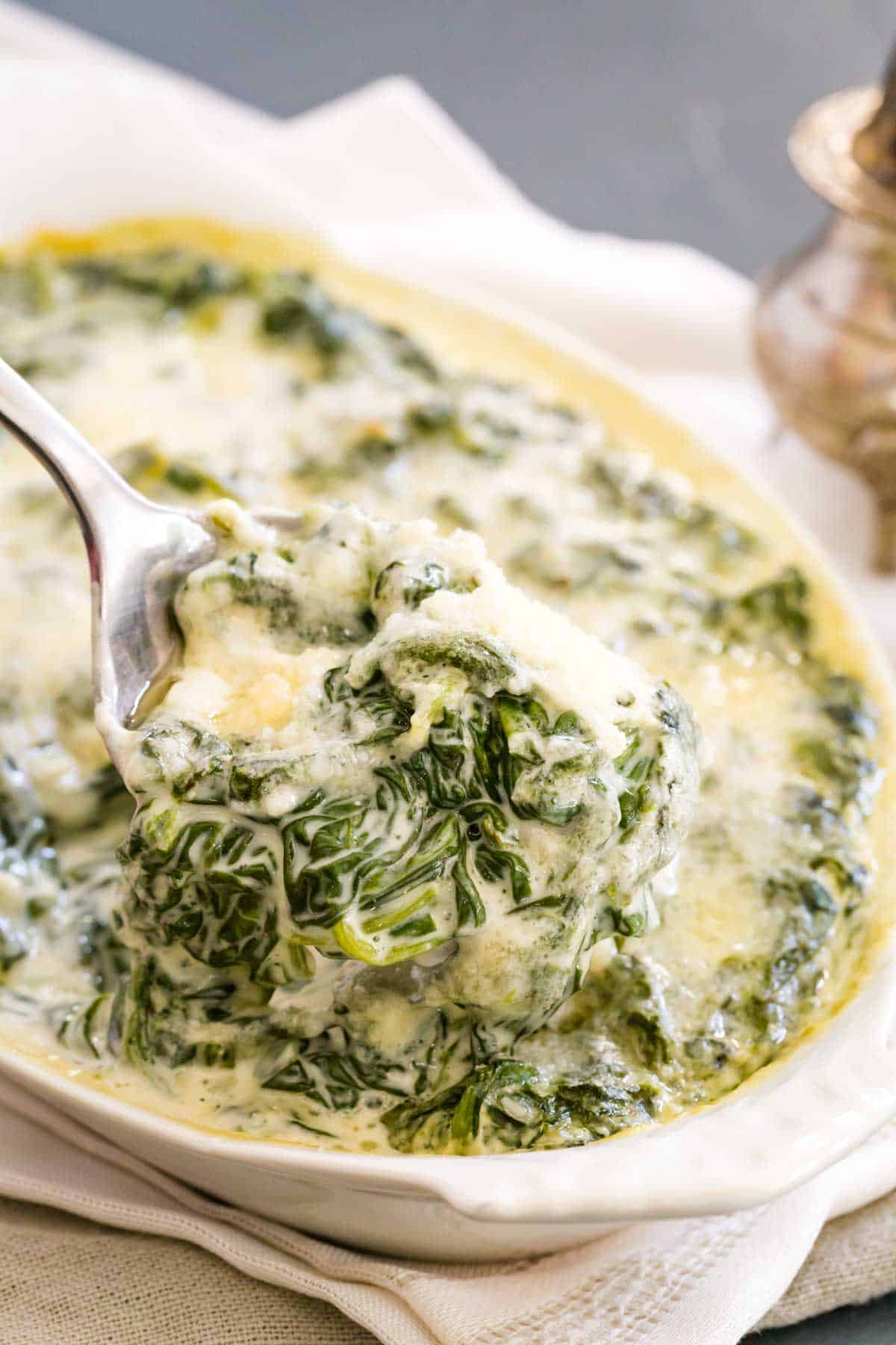A serving spoon lifts out cheese-topped cream spinach to show the cooked spinach inside.