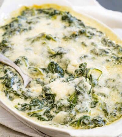 A white baking dish set on a white napkin holds creamed spinach, with a spoon digging in to scoop some out.