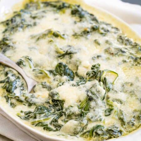 A white baking dish set on a white napkin holds creamed spinach, with a spoon digging in to scoop some out.