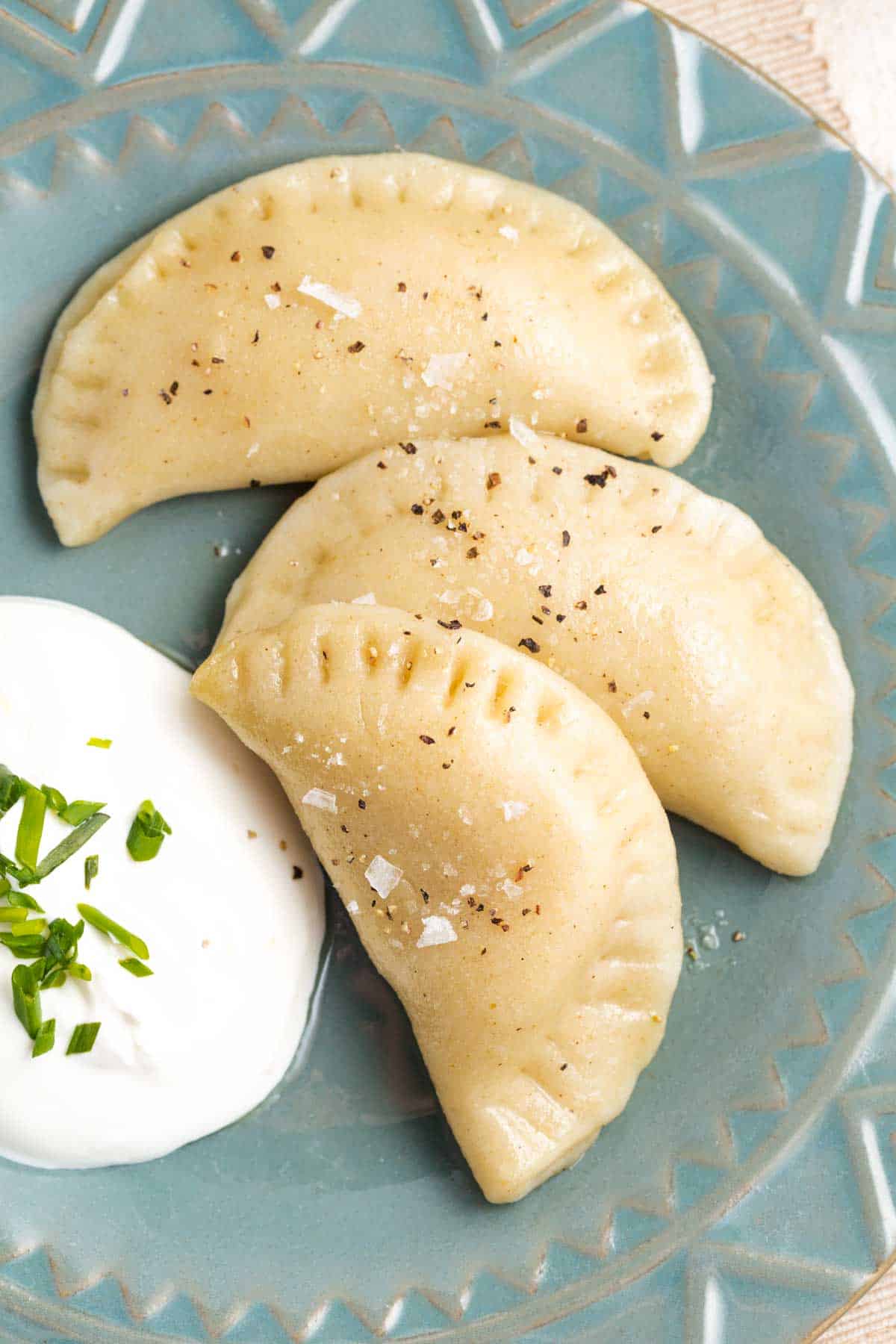 Three pierogi are shown on a blue plate, with salt and pepper visible on their tops. Next to the pierogi is a spoonful of sour cream topped with chopped parsley.