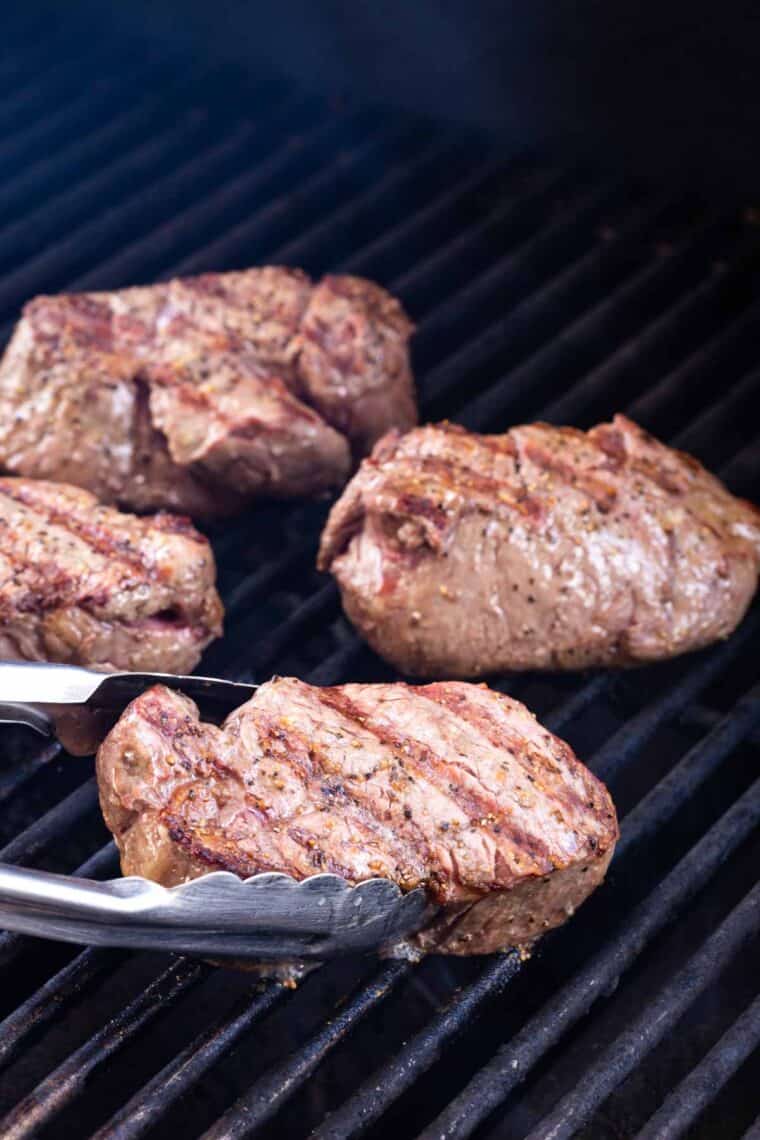 Four grilling steaks are shown on a black grill with tongs turning one of the steaks.
