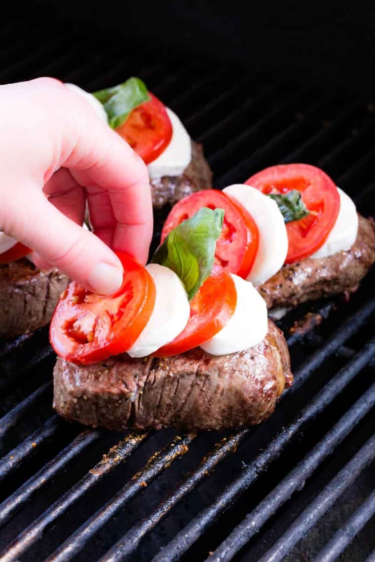 A hand layers steaks on the grill with fresh mozzarella, tomato slices, and fresh basil leaves.