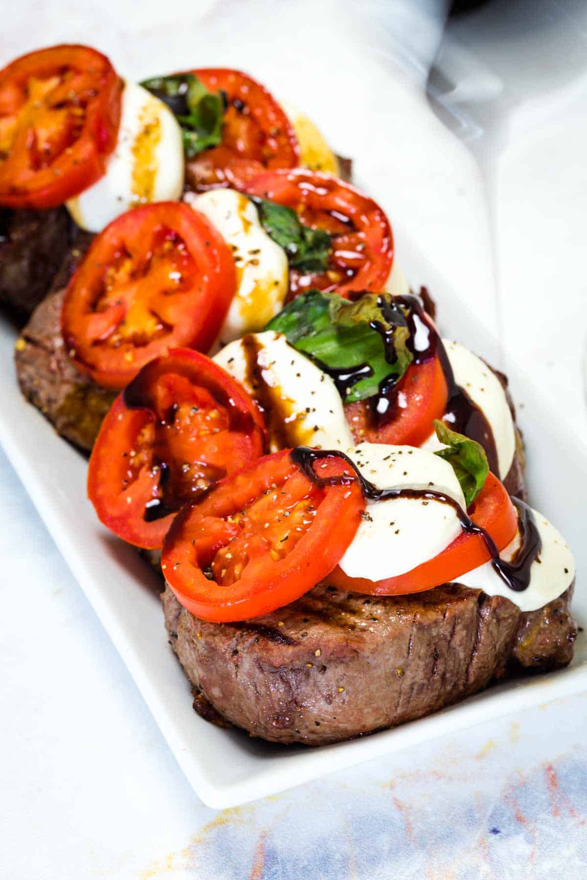 A white tray holds four colorful Caprese steaks topped with slices of fresh mozzarella, bright red tomatoes, fresh basil leaves, and a drizzle of balsamic vinegar reduction.