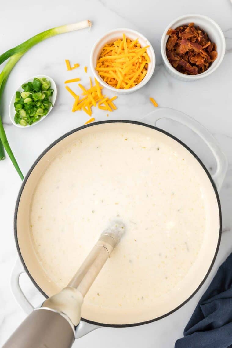 An immersion blender blends a pot of cream of potato soup, with small bowls of bacon and shredded cheese seen to the side,