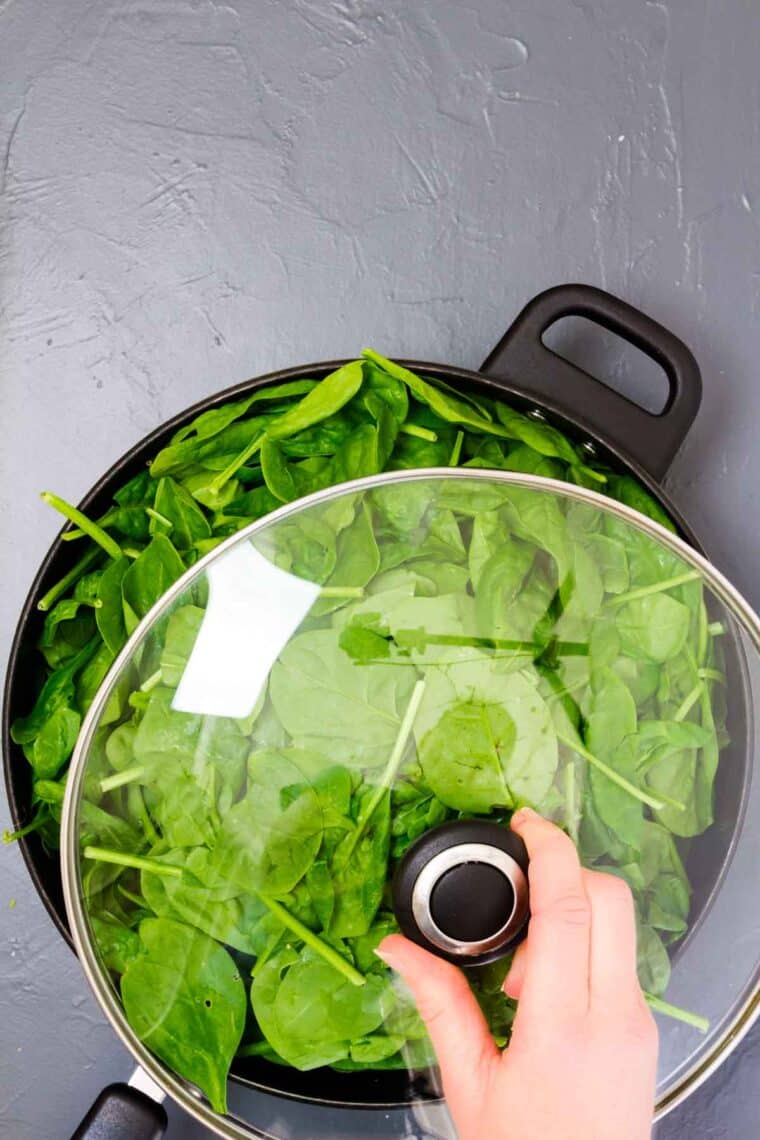 A hand places the glass cover on a black skillet full of baby spinach leaves.