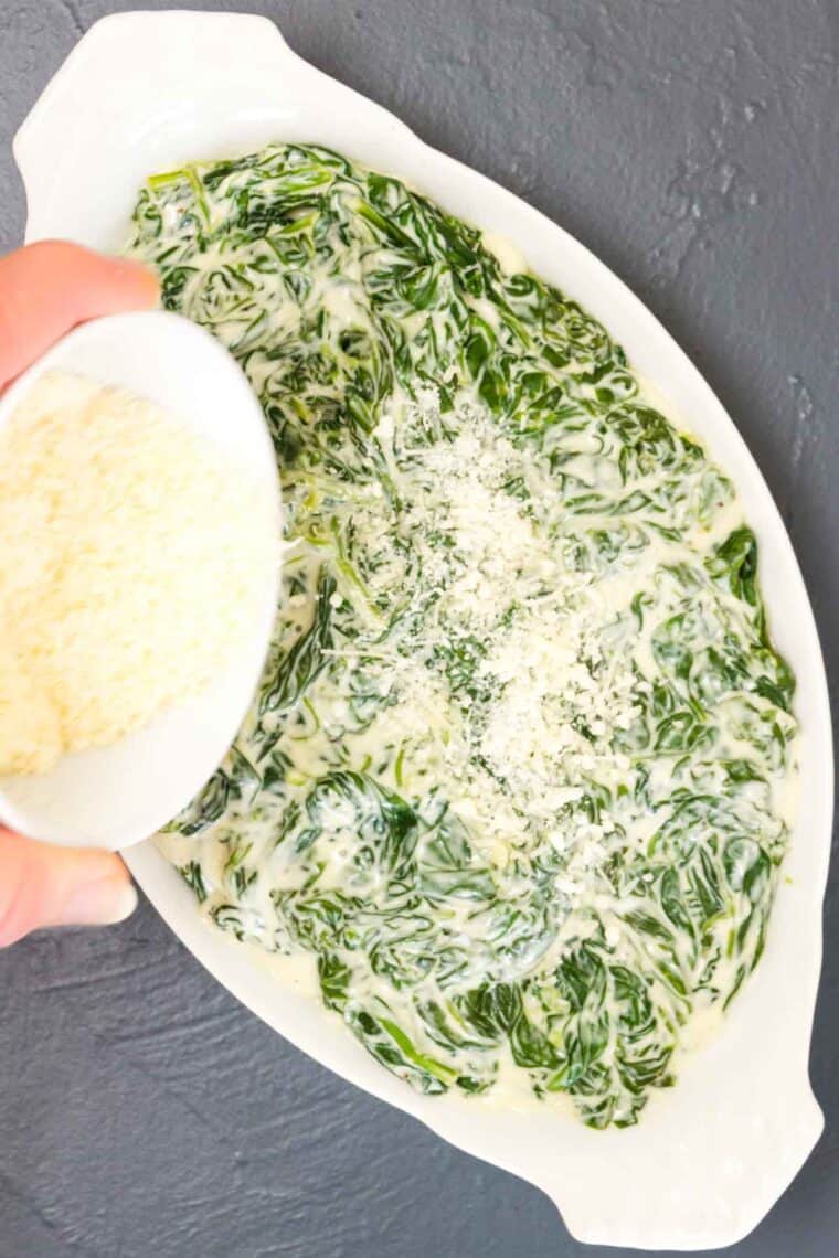 Parmesan cheese is added to a white baking pan of cooked spinach and cream cheese.
