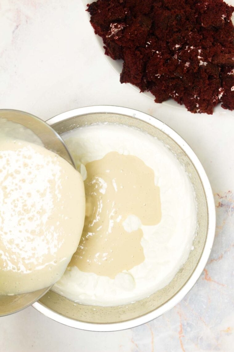 A metal bowl of ice cream base is poured into a metal bowl of whipped cream.