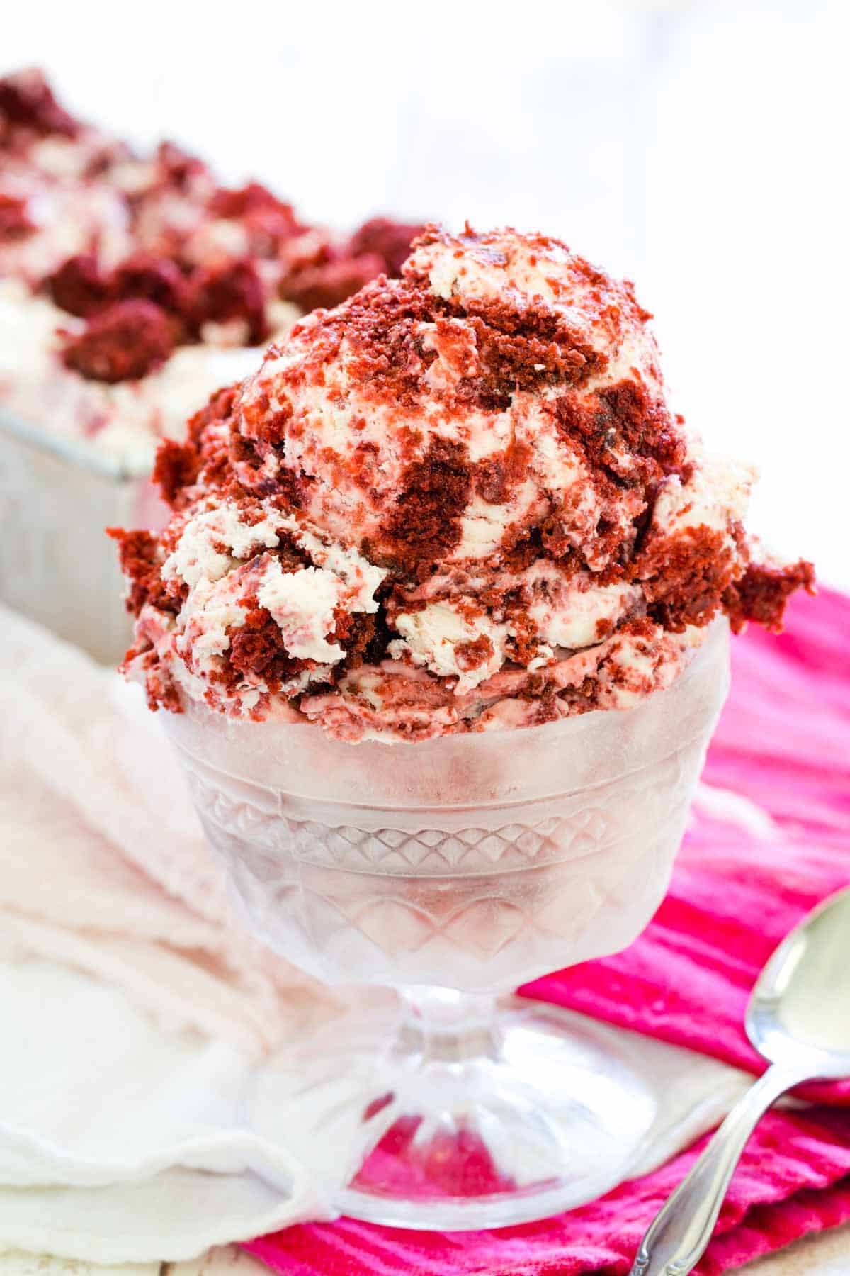 Red and white scoops of velvet ice cream are shown in a glass ice cream dish on a red napkin with a spoon next to it.