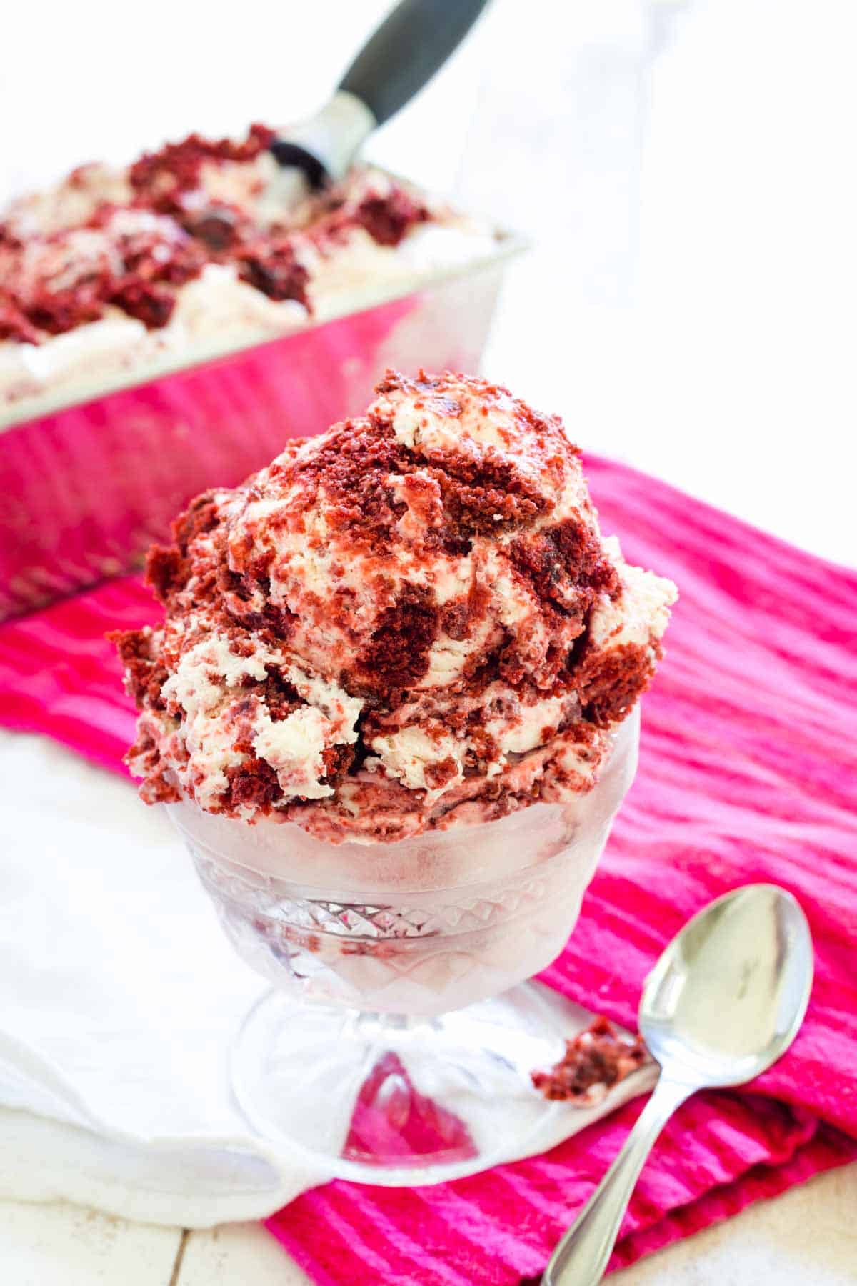 A glass ice cream dish holds a big scoop of red and white ice cream. It's placed on a red napkin alongside a spoon, and a loaf pan of red velvet ice cream with an ice cream scoop is seen in the background.