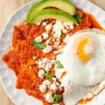 A big plate of chilaquiles rojos is shown close up, with lots of contrasting colors including red sauce, white cheese crumbles, a white and yellow fried eggs, and green parsley.