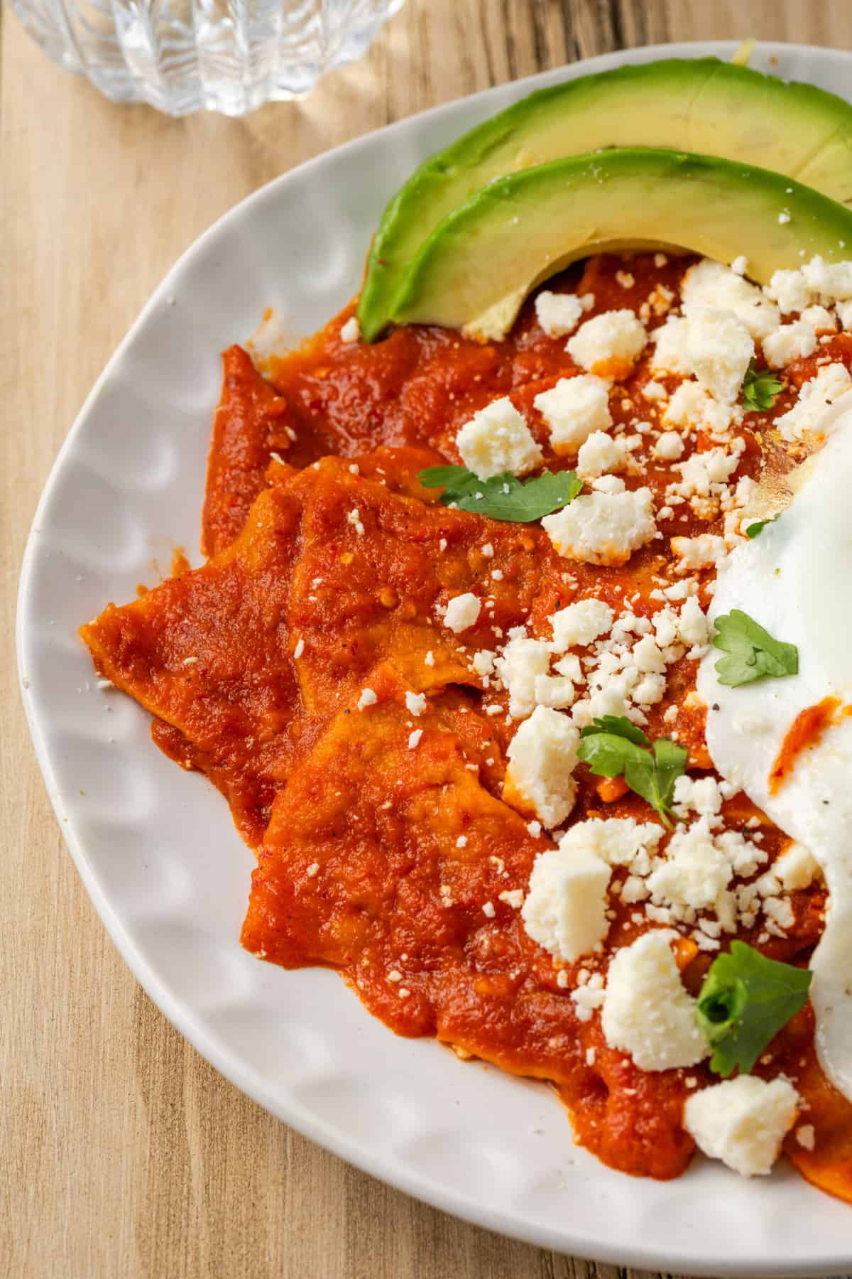 Chilaquiles rojos are shown on a white plate topped with red sauce, crumbled cheese, parsley, sliced avocado, and a fried egg.