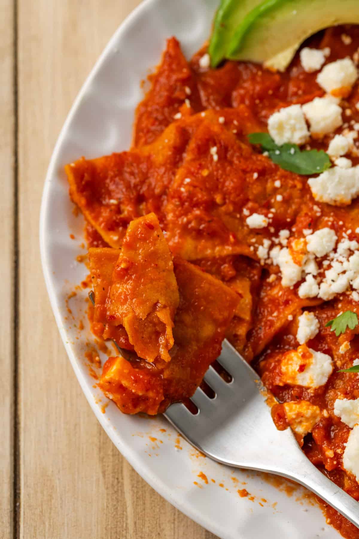 A fork cuts into a plate of chilaquiles rojos, shown in a half-view that features the red sauce.