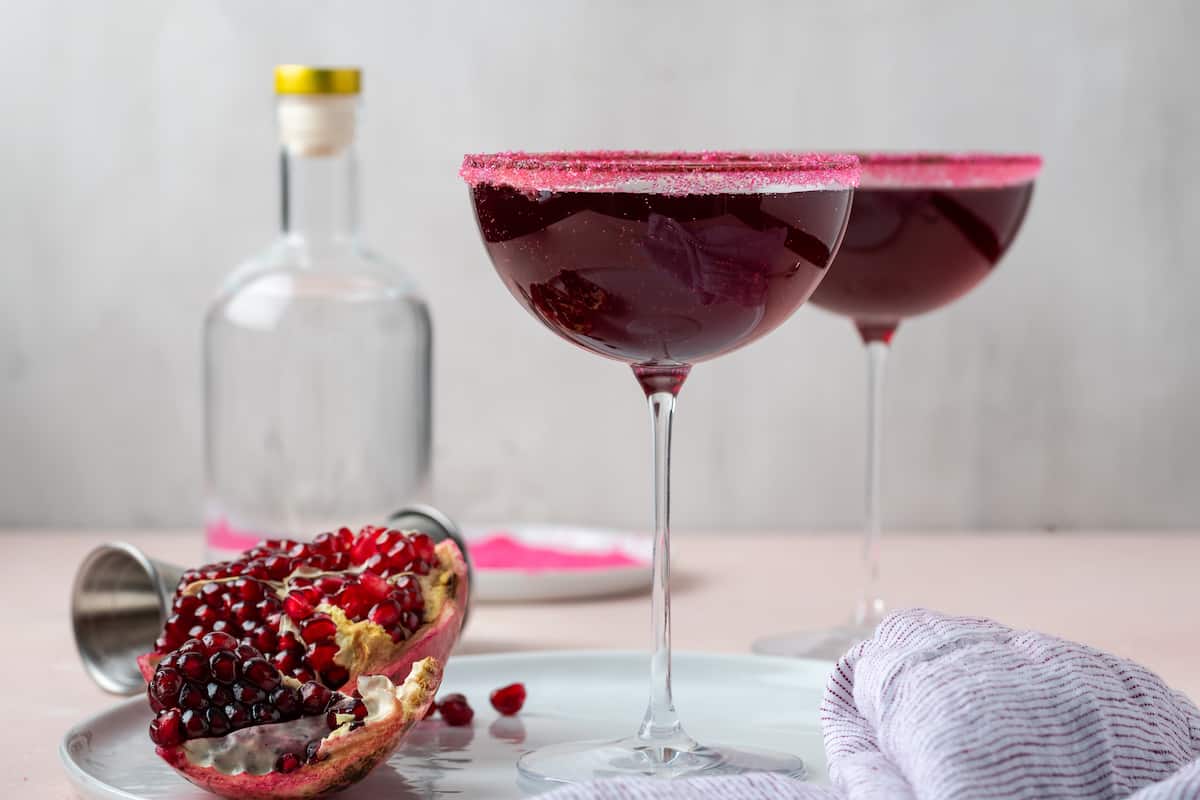Two tall coupe glasses rimmed with bright pink sugar stand next to each other with a bottle in the background and a cut pomegranate nearby.