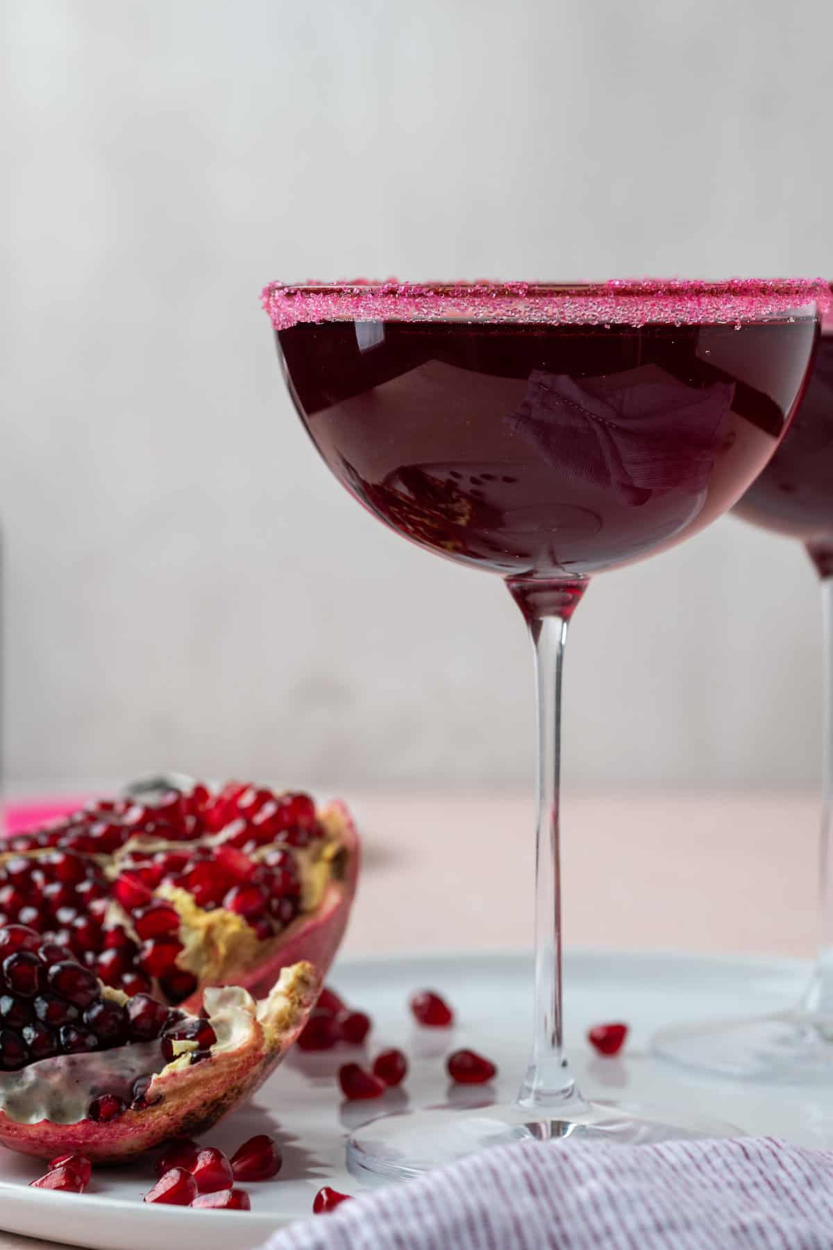 A tall coupe glass of pomegranate cocktail stands next to a cut open pomegranate that shows its seeds.