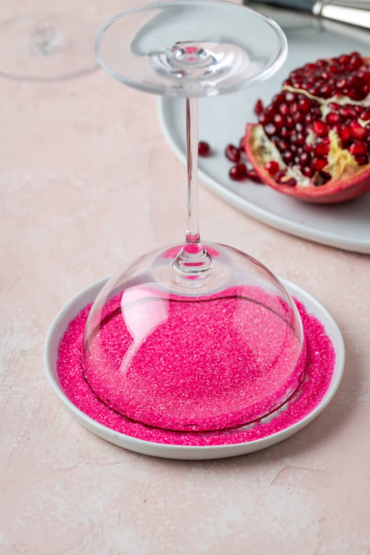 The rim of a glass cocktail glass is dipped into a small bowl of neon pink sugar.
