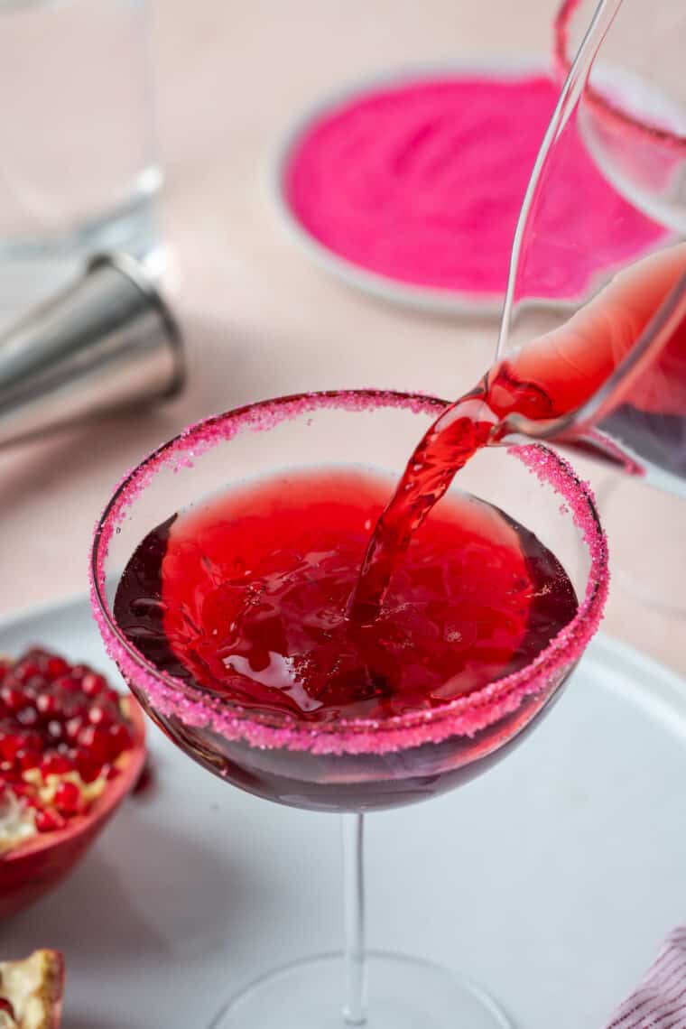 A glass pitched pours dark red pomegranate cocktail into a glass that's rimmed with pink sugar.