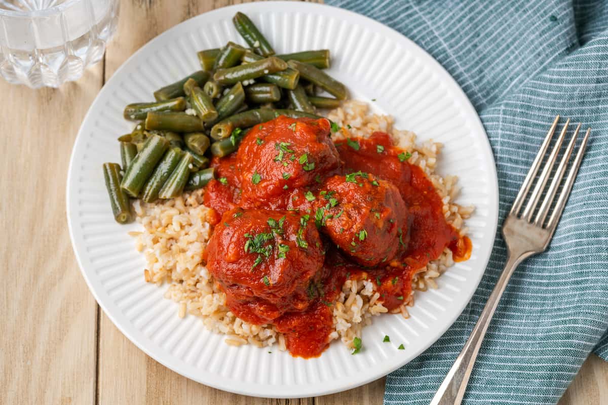 A white plate holds porcupine meatballs in red sauce alongside green beans, with rice.