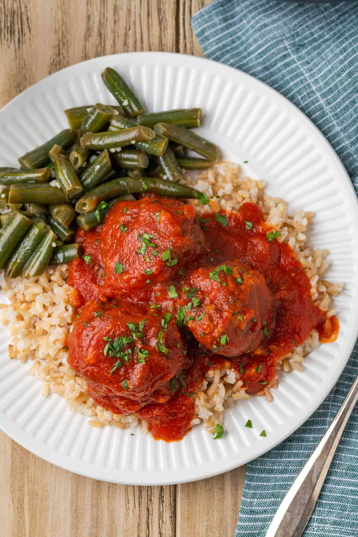 Porcupine meatballs served over rice with green beans are shown on a white plate with a fork and blue napkin to the right.