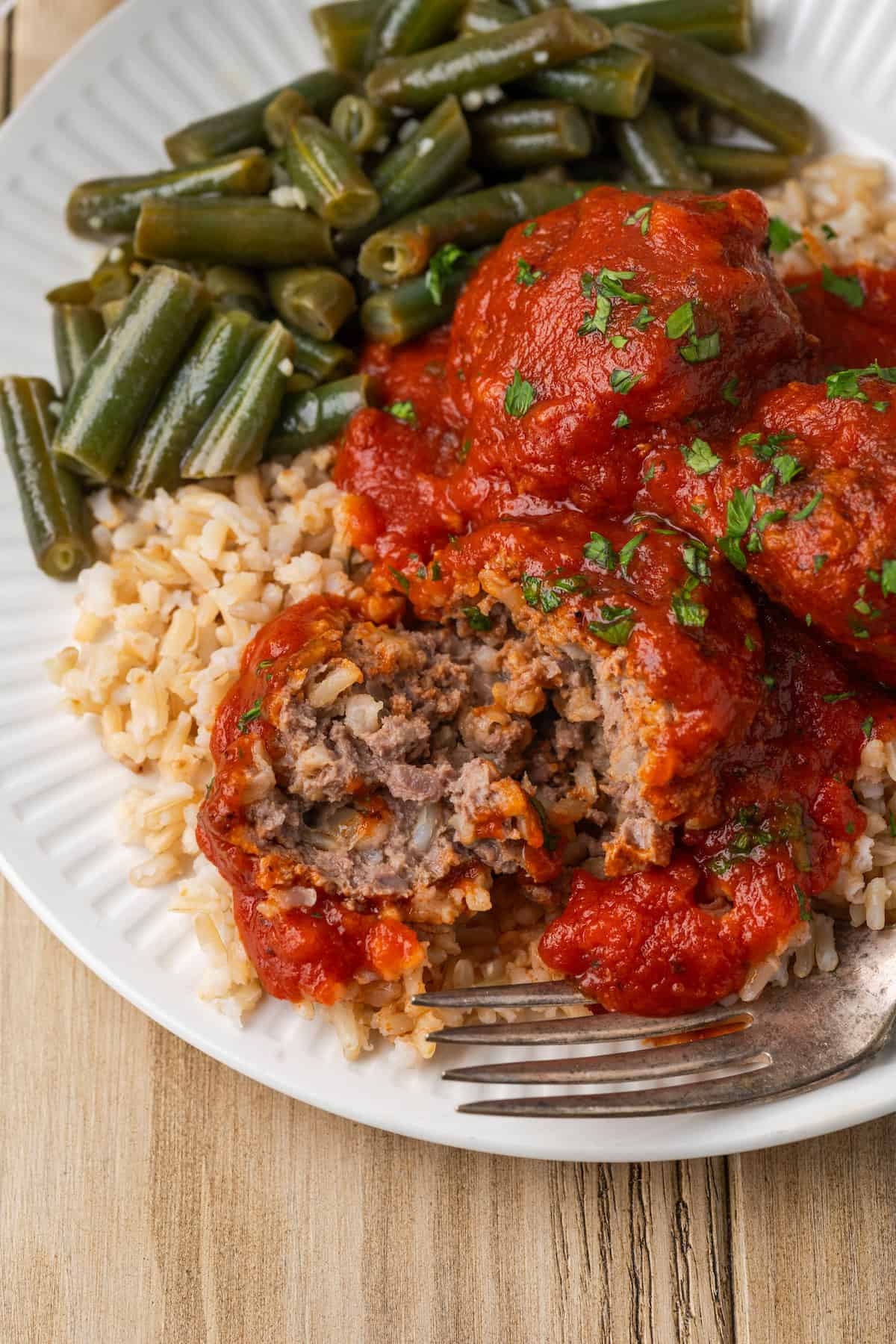 A cut porcupine meatball shows its meaty interior. It has a fork next to it and is served in red sauce over rice and green beans.