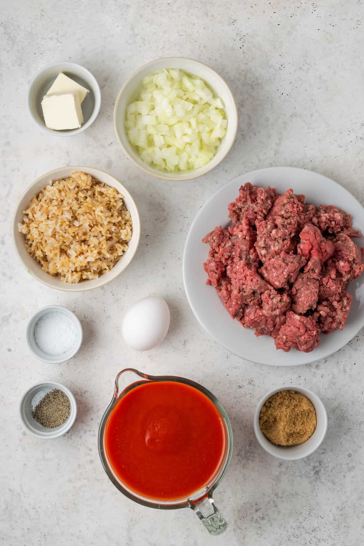 The ingredients needed to make porcupine meatballs are shown portioned out on a white background: ground beef, chopped onions, brown rice, salt and pepper, tomato sauce, egg, and butter.