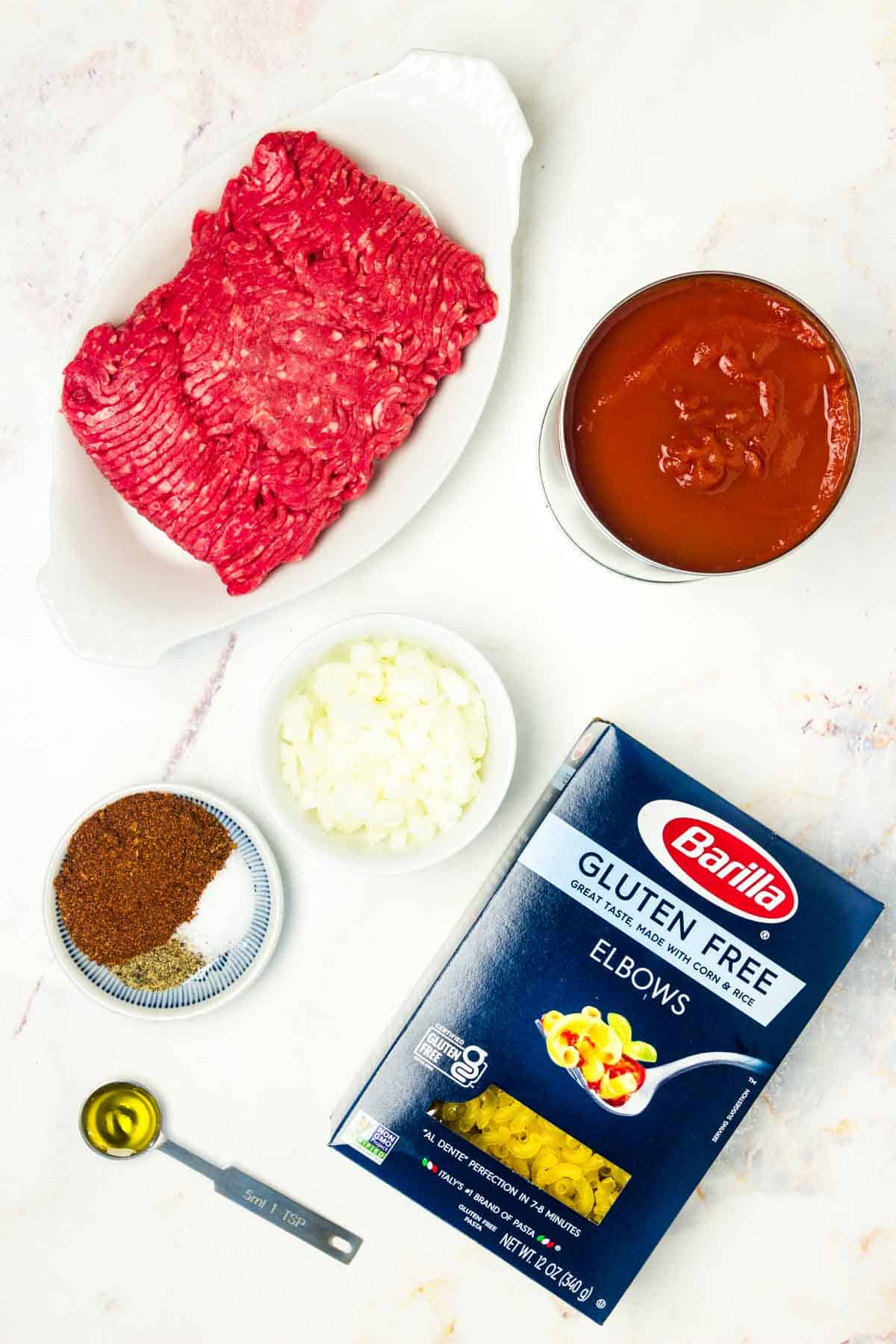 The ingredients for beefaroni are shown portioned out: ground beef, onion, garlic, gluten free elbows, tomato sauce, olive oil, chili powder, salt and pepper.
