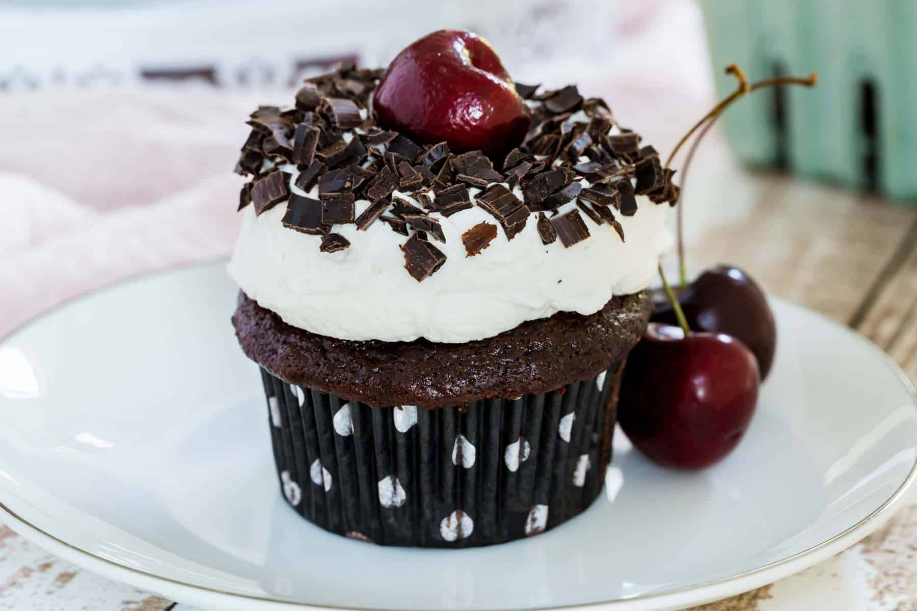 A black forest cupcake with a cherry next to it is on a white plate.