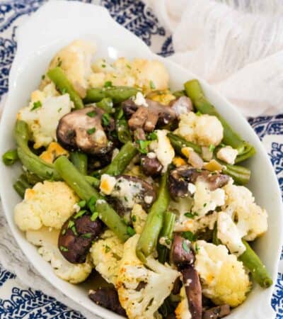 A white serving dish on a blue and white napkin holds a pile of blue cheese roasted vegetables.