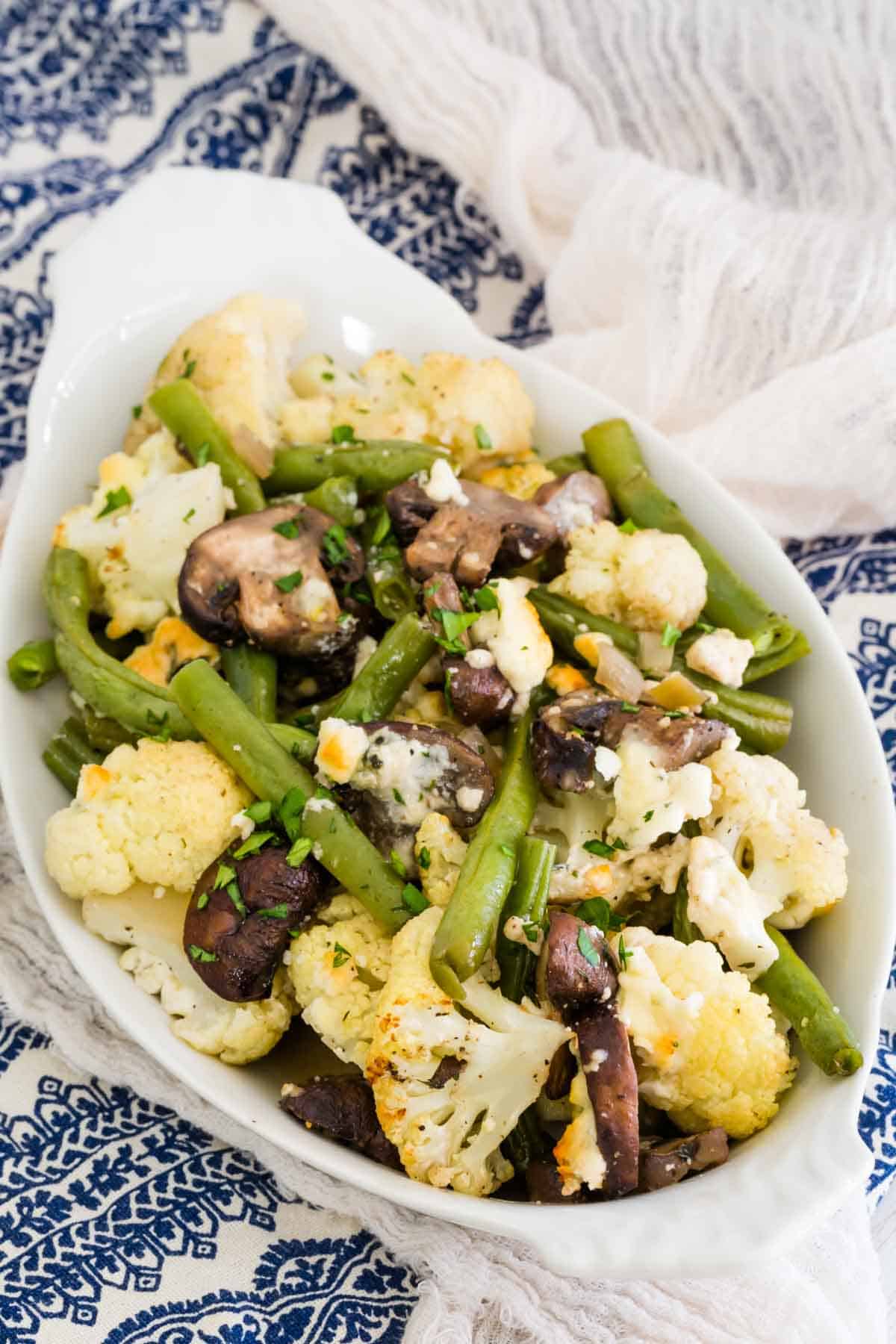 A white serving dish on a blue and white napkin holds a pile of blue cheese roasted vegetables.