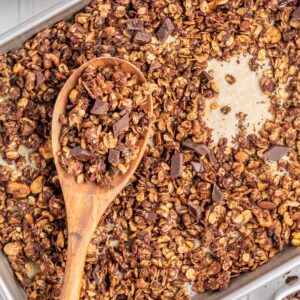 A wooden spoon resting on a sheet pan of chocolate granola.