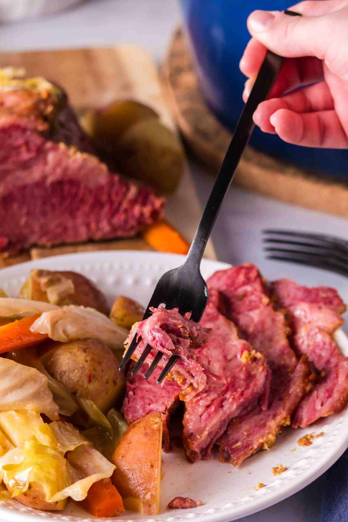 A hand holding a fork digs into a plate of corned beef with cabbage and potatoes.