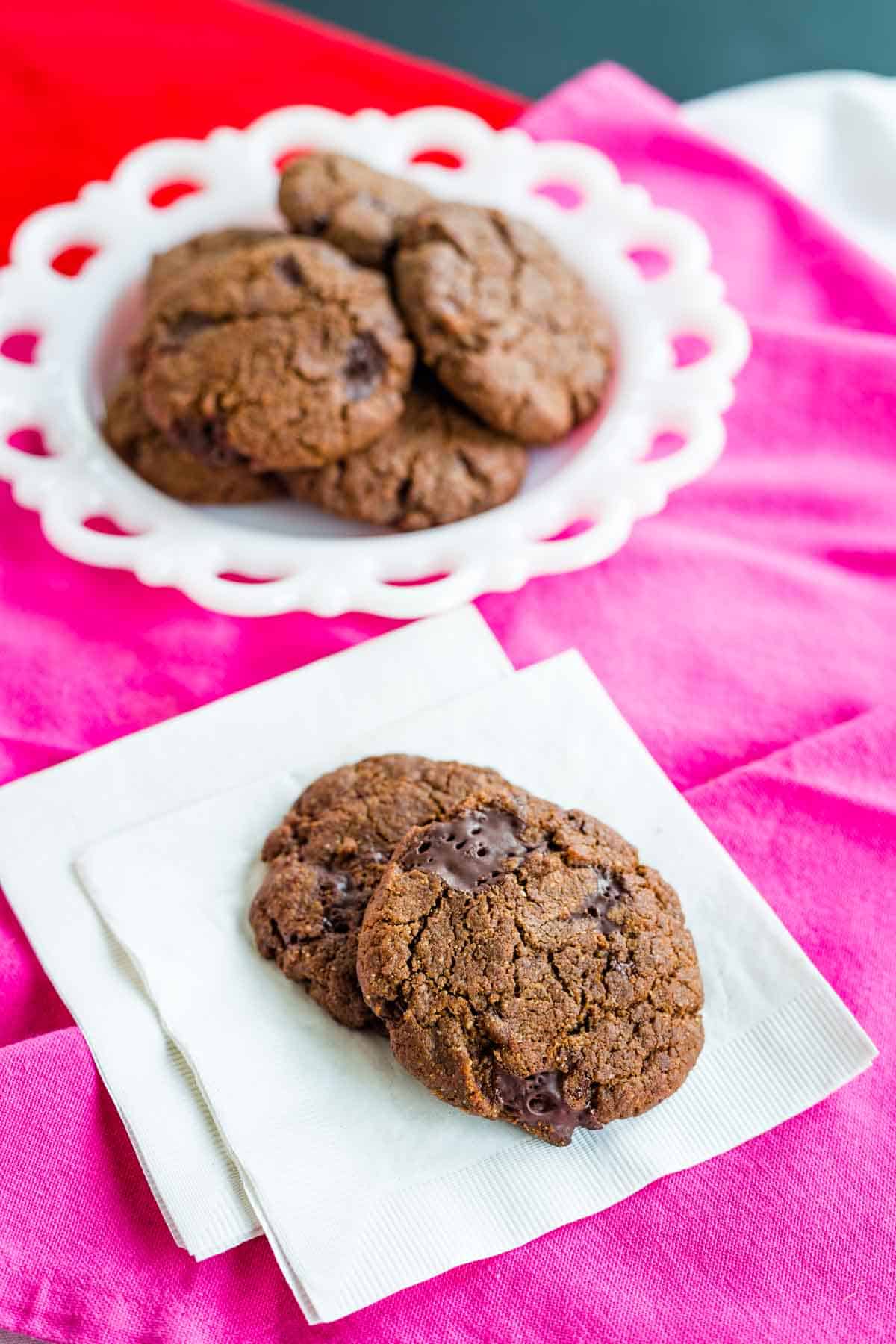 Two dark chocolate chunk cookies are shown on napkins with a plate of cookies behind them on a pink tablecloth.