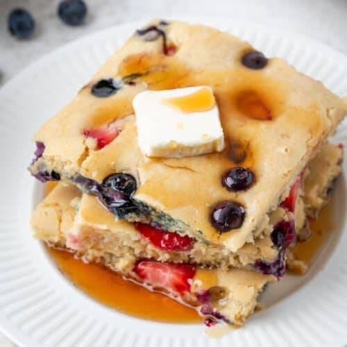 A stack of three pieces of sheet pan pancakes filled with berries on a plate with butter and syrup.
