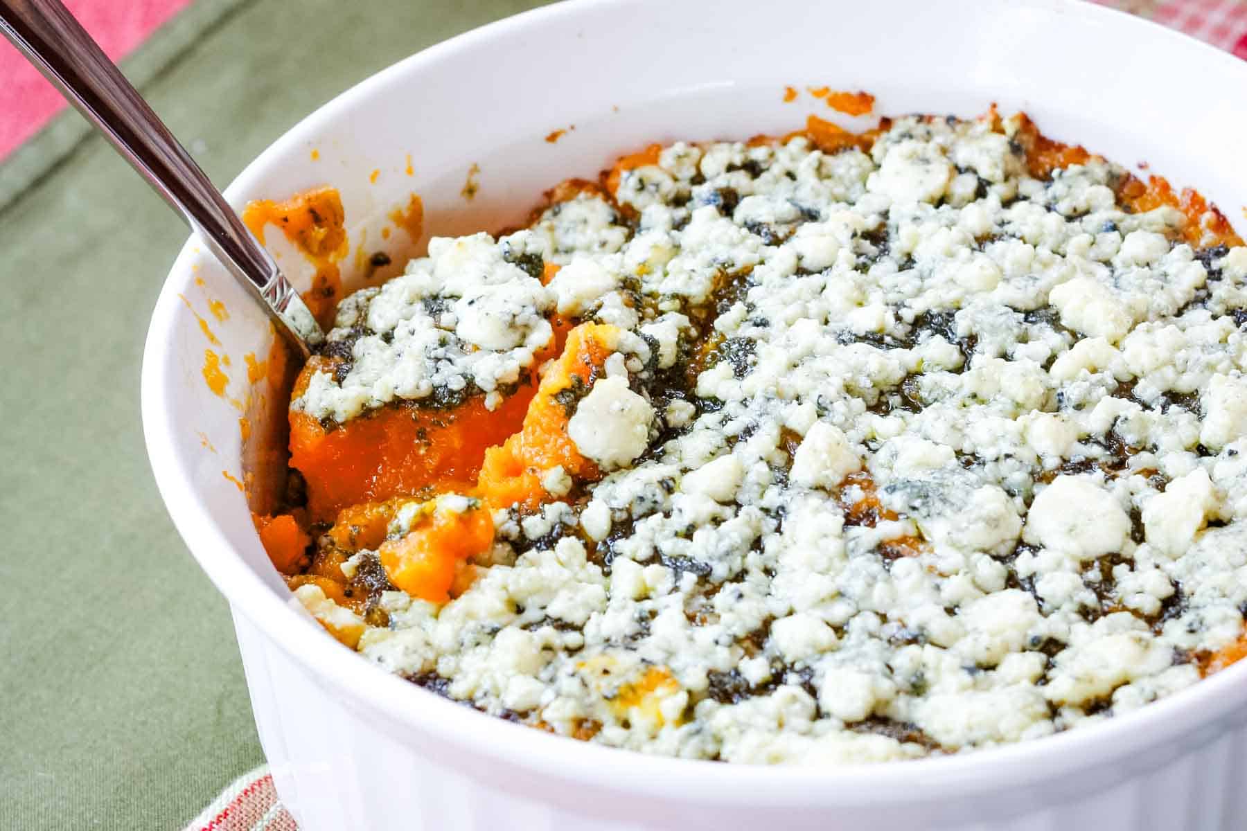 A spoon in a casserole dish of Butternut Squash Gratin topped with gorgonzola cheese.